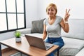 Middle age blonde woman using laptop at home smiling with happy face winking at the camera doing victory sign with fingers Royalty Free Stock Photo