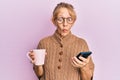 Middle age blonde woman using smartphone and drinking a cup of coffee making fish face with mouth and squinting eyes, crazy and Royalty Free Stock Photo