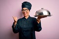 Middle age brunette chef woman wearing cooker uniform and hat holding tray dome pointing and showing with thumb up to the side Royalty Free Stock Photo