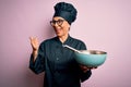 Middle age brunette chef woman wearing cooker uniform and hat using whisk and bowl pointing and showing with thumb up to the side Royalty Free Stock Photo