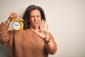 Middle age brunette woman holding clasic alarm clock over isolated background doing stop gesture with hands palms, angry and Royalty Free Stock Photo