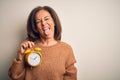 Middle age brunette woman holding clasic alarm clock over isolated background sticking tongue out happy with funny expression Royalty Free Stock Photo