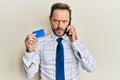 Middle age businessman talking on the smartphone holding credit card in shock face, looking skeptical and sarcastic, surprised