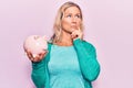 Middle age caucasian blonde woman holding piggy bank serious face thinking about question with hand on chin, thoughtful about Royalty Free Stock Photo