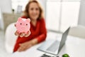 Middle age caucasian woman holding piggy bank sitting on the table at home Royalty Free Stock Photo