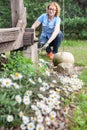 Middle age Caucasian woman sits with gloves near wooden veranda and cares for camomiles growing in backyard