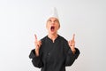 Middle age chef woman wearing uniform and hat standing over isolated white background amazed and surprised looking up and pointing Royalty Free Stock Photo