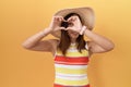 Middle age chinese woman wearing summer hat over yellow background doing heart shape with hand and fingers smiling looking through Royalty Free Stock Photo