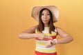 Middle age chinese woman wearing summer hat over yellow background in hurry pointing to watch time, impatience, upset and angry