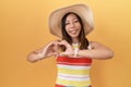 Middle age chinese woman wearing summer hat over yellow background smiling in love doing heart symbol shape with hands Royalty Free Stock Photo