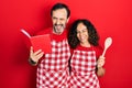 Middle age couple of hispanic woman and man wearing professional apron reading cooking recipe book winking looking at the camera Royalty Free Stock Photo