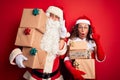 Middle age couple wearing Santa costume holding tower of gifts over isolated red background pointing finger up with successful