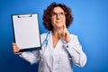 Middle age curly hair doctor woman wearing coat and stethoscope holding clipboard serious face thinking about question, very