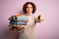 Middle age curly hair housework woman holding pile of clothes over isolated pink background pointing with finger to the camera and