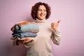 Middle age curly hair housework woman holding pile of clothes over isolated pink background pointing and showing with thumb up to