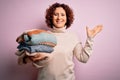Middle age curly hair housework woman holding pile of clothes over isolated pink background very happy and excited, winner