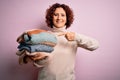 Middle age curly hair housework woman holding pile of clothes over isolated pink background very happy pointing with hand and