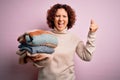 Middle age curly hair housework woman holding pile of clothes over pink background screaming proud and celebrating