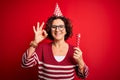 Middle age curly hair woman wearing birthday funny hat holding party trumpet on celebration doing ok sign with fingers, excellent Royalty Free Stock Photo