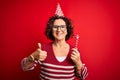 Middle age curly hair woman wearing birthday funny hat holding party trumpet on celebration happy with big smile doing ok sign, Royalty Free Stock Photo