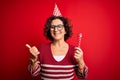Middle age curly hair woman wearing birthday funny hat holding party trumpet on celebration pointing and showing with thumb up to Royalty Free Stock Photo