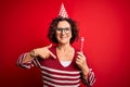 Middle age curly hair woman wearing birthday funny hat holding party trumpet on celebration with surprise face pointing finger to Royalty Free Stock Photo