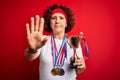 Middle age curly woman winning medals holding trophy over isolated red background with open hand doing stop sign with serious and