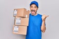 Middle age delivery man wearing cap holding package over isolated white background pointing thumb up to the side smiling happy Royalty Free Stock Photo