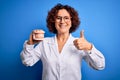 Middle age dentist woman wearing coat holding plastic denture teeth over blue background happy with big smile doing ok sign, thumb Royalty Free Stock Photo