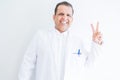 Middle age doctor man wearing medical coat over white background smiling with happy face winking at the camera doing victory sign Royalty Free Stock Photo