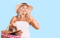 Middle age fit blonde woman wearing summer hat and holding picnic wicker basket with bread smiling happy and positive, thumb up