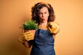 Middle age gardener woman wearing apron holding plant pot over isolated yellow background pointing with finger to the camera and