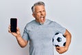 Middle age grey-haired man holding football ball showing smartphone angry and mad screaming frustrated and furious, shouting with Royalty Free Stock Photo