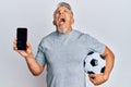 Middle age grey-haired man holding football ball showing smartphone angry and mad screaming frustrated and furious, shouting with Royalty Free Stock Photo