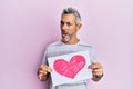Middle age grey-haired man holding heart draw in shock face, looking skeptical and sarcastic, surprised with open mouth Royalty Free Stock Photo