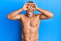 Middle age grey-haired man standing shirtless doing ok gesture like binoculars sticking tongue out, eyes looking through fingers