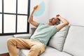 Middle age grey-haired man using handfan sitting on sofa at home Royalty Free Stock Photo