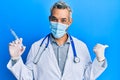 Middle age grey-haired man wearing doctor uniform and medical mask holding syringe pointing thumb up to the side smiling happy Royalty Free Stock Photo