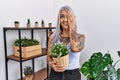 Middle age grey-haired woman holding green plant pot at home covering one eye with hand, confident smile on face and surprise Royalty Free Stock Photo