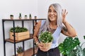 Middle age grey-haired woman holding green plant pot at home smiling with hand over ear listening an hearing to rumor or gossip Royalty Free Stock Photo