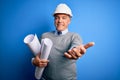 Middle age handsome grey-haired architect man wearing safety helmet holding blueprints smiling cheerful offering palm hand giving