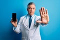 Middle age handsome grey-haired doctor man holding smartphone showing screen with open hand doing stop sign with serious and Royalty Free Stock Photo