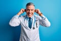 Middle age handsome grey-haired doctor man wearing coat and blue stethoscope Smiling pulling ears with fingers, funny gesture