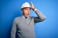 Middle age handsome grey-haired engineer man wearing safety helmet over blue background surprised with hand on head for mistake,