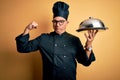 Middle age handsome grey-haired waiter man wearing cooker uniform and hat holding tray Strong person showing arm muscle, confident