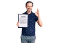 Middle age handsome man holding clipboard with contract document doing ok sign with fingers, smiling friendly gesturing excellent