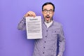 Middle age handsome man wearing glasses holding contract document over purple background scared and amazed with open mouth for Royalty Free Stock Photo