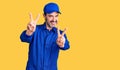 Middle age handsome man wearing mechanic uniform smiling with tongue out showing fingers of both hands doing victory sign Royalty Free Stock Photo