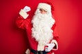 Middle age handsome man wearing Santa costume standing over isolated red background Strong person showing arm muscle, confident Royalty Free Stock Photo