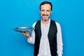 Middle age handsome waitress man holding tray standing over isolated blue background looking positive and happy standing and